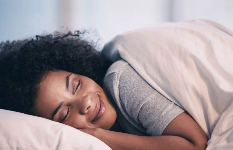 Sleeping, woman and smile in bed with morning nap at home with rest feeling calm with peace. House, bedroom and tired female person relax and comfortable on a pillow with blanket dreaming on weekend | Amarillo Medical Specialists Rheumatology
